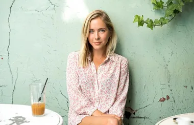 A woman in a floral shirt sits against a green wall, with a glass of iced coffee on a white table beside her.