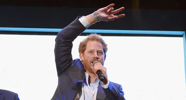 Man in a suit speaking into a microphone, raising one arm with fingers in a peace symbol.
