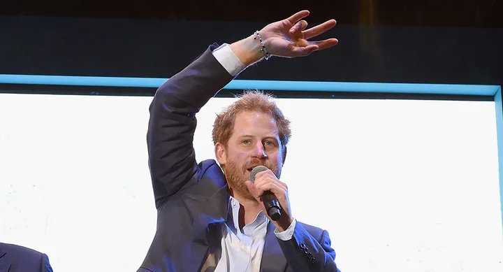 Man in a suit speaking into a microphone, raising one arm with fingers in a peace symbol.