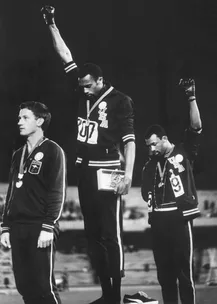 Three athletes on a podium; two raise fists with Olympic medals, one stands solemn; iconic protest moment.
