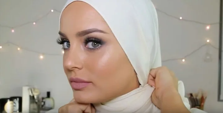 Woman wearing a white hijab, applying makeup in a softly lit room with fairy lights in the background.