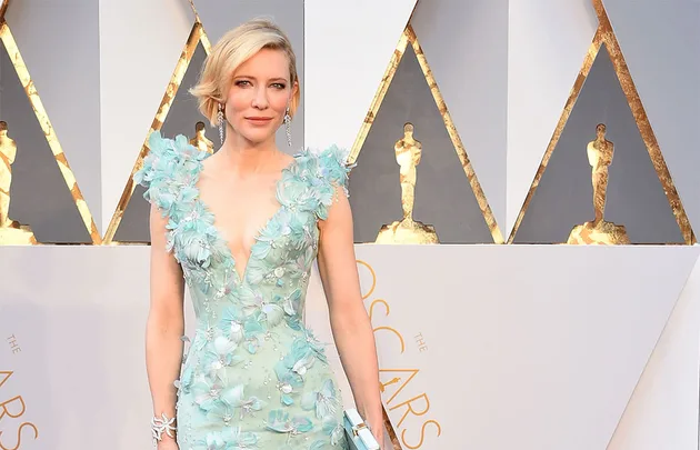 Woman in a light blue floral gown at an awards event with gold Oscar statuette decor in the background.