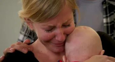 A woman cries while holding a baby close, with a comforting hand on her shoulder.
