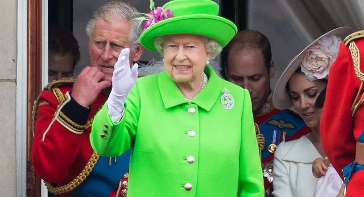 A woman in a bright green outfit waves from a balcony, surrounded by others in formal attire.