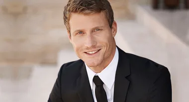 A person in a suit and tie smiling, sitting outdoors against a blurred background.