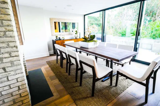 Dining room with a large wooden table, white chairs, and a large window overlooking greenery.