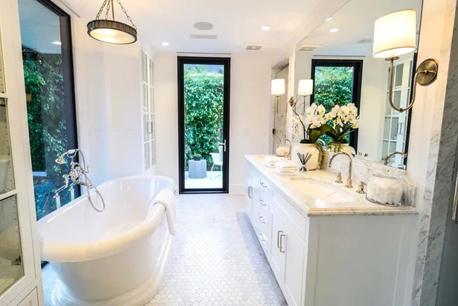 Luxurious bathroom with a freestanding tub, double sink vanity, floral decor, and views of greenery through large windows.