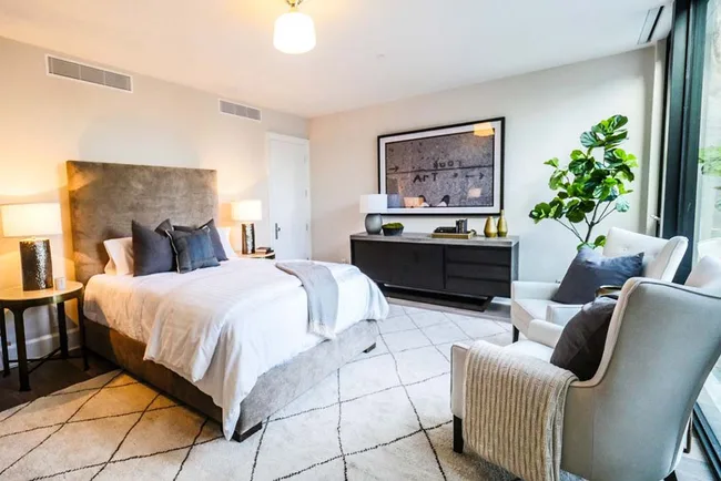 Contemporary bedroom with a large bed, cushioned headboard, two side tables, an armchair, artwork, and a potted plant.