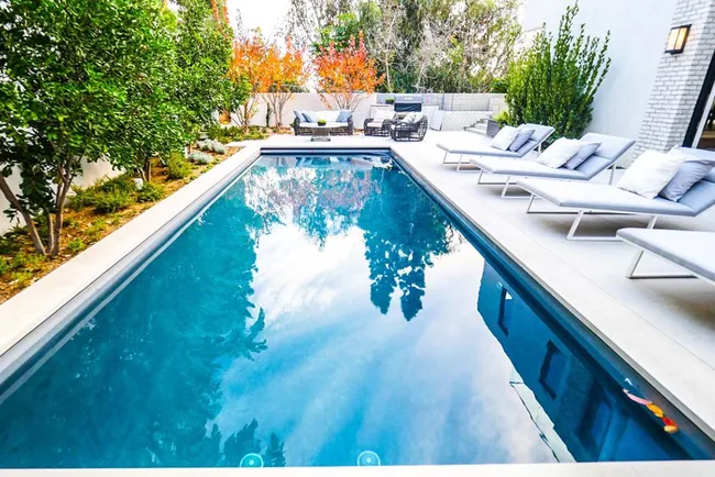 Modern backyard pool with lounge chairs, surrounded by trees and autumn foliage, reflecting blue sky and clouds.