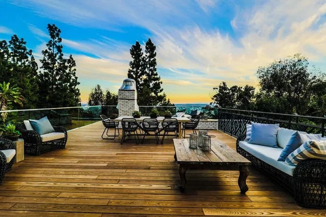Outdoor wooden deck with seating, a large table, and a scenic view of trees and sky at sunset.