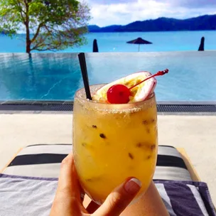 A tropical cocktail with a cherry and straw, held by a pool with a view of the ocean and distant hills.