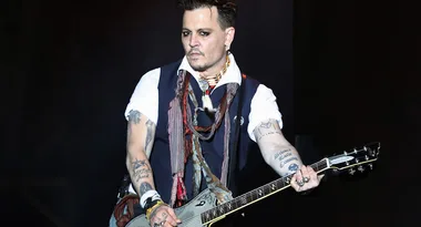 A person playing an electric guitar on stage, wearing a vest and scarf, with visible tattoos and jewelry.