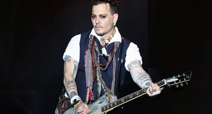 A person playing an electric guitar on stage, wearing a vest and scarf, with visible tattoos and jewelry.