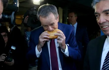 Man in a suit eating a sandwich while cameras flash around him.