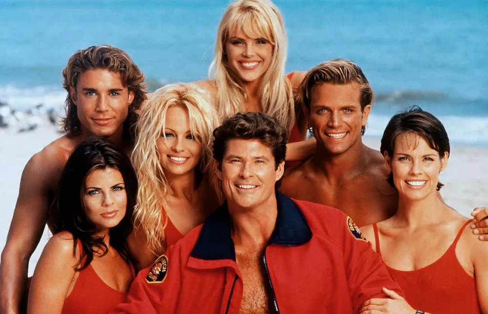 Group of lifeguards in red swimwear and jackets on a beach, smiling towards the camera.