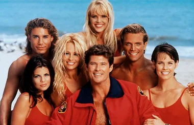 Group of lifeguards in red swimwear and jackets on a beach, smiling towards the camera.