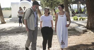 Three people walking and smiling outdoors on a sunny day, with trees and a white building in the background.