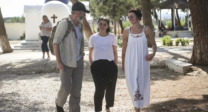 Three people walking and smiling outdoors on a sunny day, with trees and a white building in the background.