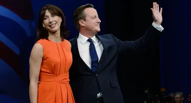 Couple standing together, the woman in an orange dress smiles, while the man in a suit waves to a crowd.