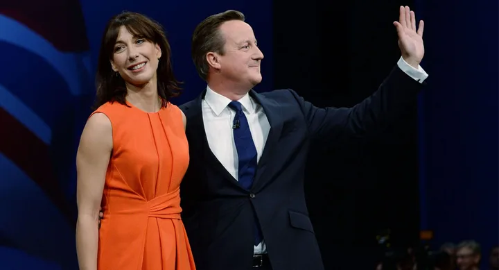 Couple standing together, the woman in an orange dress smiles, while the man in a suit waves to a crowd.