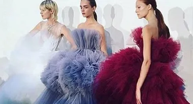 Models wearing elaborate, voluminous dresses in white, blue, and burgundy on a runway.