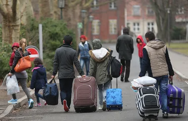 People walking down a street with luggage, dressed for cool weather, suggesting travel or relocation.