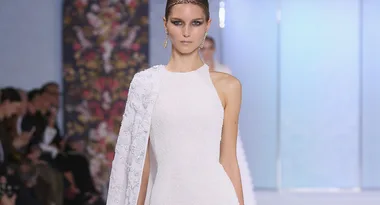 Model in white gown with floral detail on runway, floral backdrop, spectators in background.