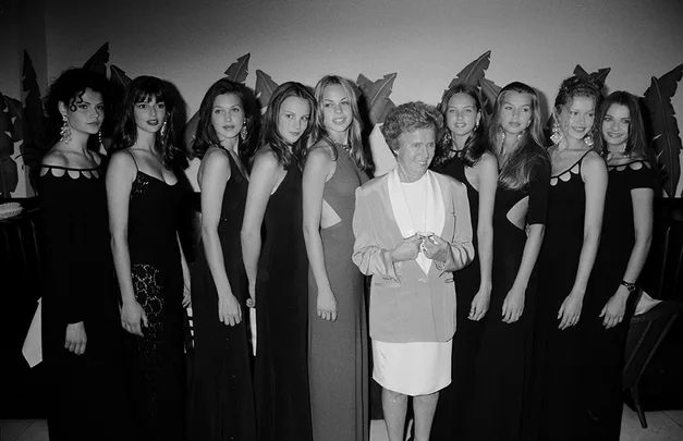 A group of women in elegant evening gowns pose together, with one older woman in a suit, indoors.