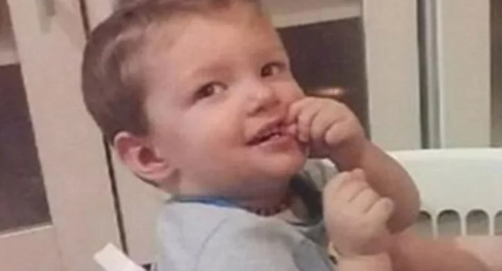 A smiling toddler sitting indoors, wearing a light blue shirt, with hands near their face.
