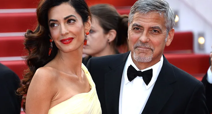 A couple poses elegantly on a red carpet; the woman wears a yellow gown and the man a black tuxedo.