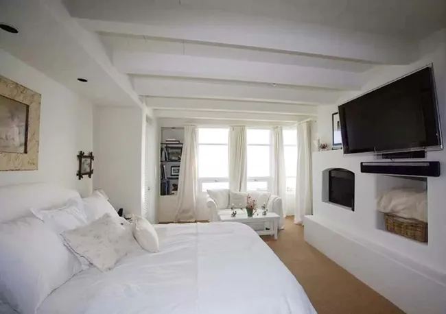 Cozy bedroom with white decor, bed, wall-mounted TV, sofa, and bookshelf by large windows with light curtains.