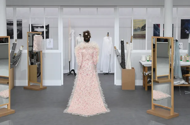 Woman in a pink floral gown with fur trim stands in a fashion atelier, surrounded by mirrors and fabric rolls.