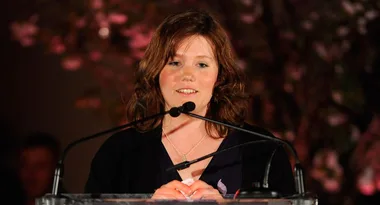 A woman with brown hair speaks into a microphone on a podium, set against a blurred background.
