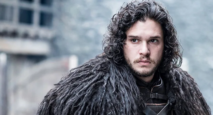 A person with dark, curly hair and a serious expression, wearing a fur cloak, stands outdoors with a blurred background.