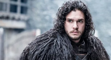 A person with dark, curly hair and a serious expression, wearing a fur cloak, stands outdoors with a blurred background.
