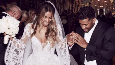 A bride in a white lace wedding dress holds hands with a groom in a black suit as they smile, surrounded by guests.