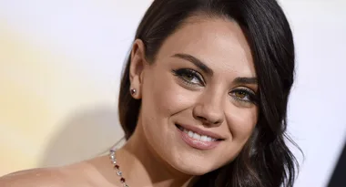 A woman with long dark hair and a smile, wearing a necklace, against a blurred background.