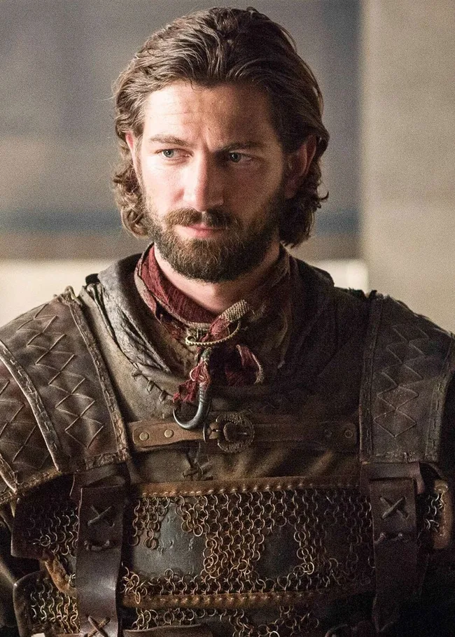 A man with a beard wearing medieval armor looks to the side in a dimly lit room.
