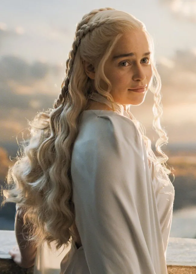 A woman with long, braided blonde hair in a white gown, looking back softly, with a blurred sunset background.