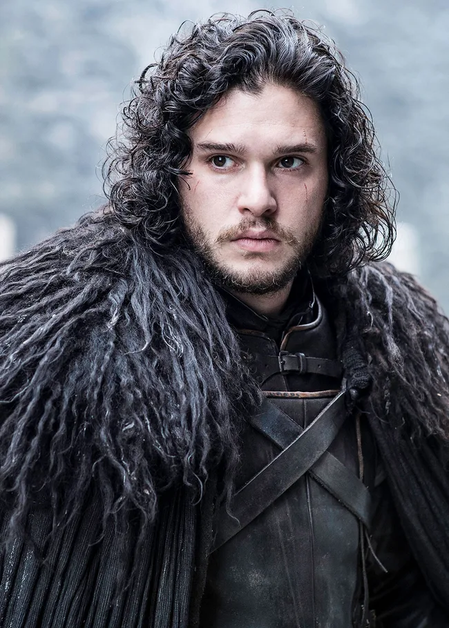 A man with curly hair and a serious expression wears a dark, fur-lined cloak against a blurred outdoor background.