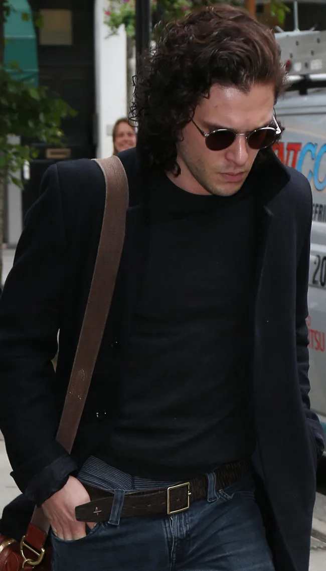 Young man with curly hair, wearing sunglasses, black coat, and jeans, walking outdoors with a shoulder bag.