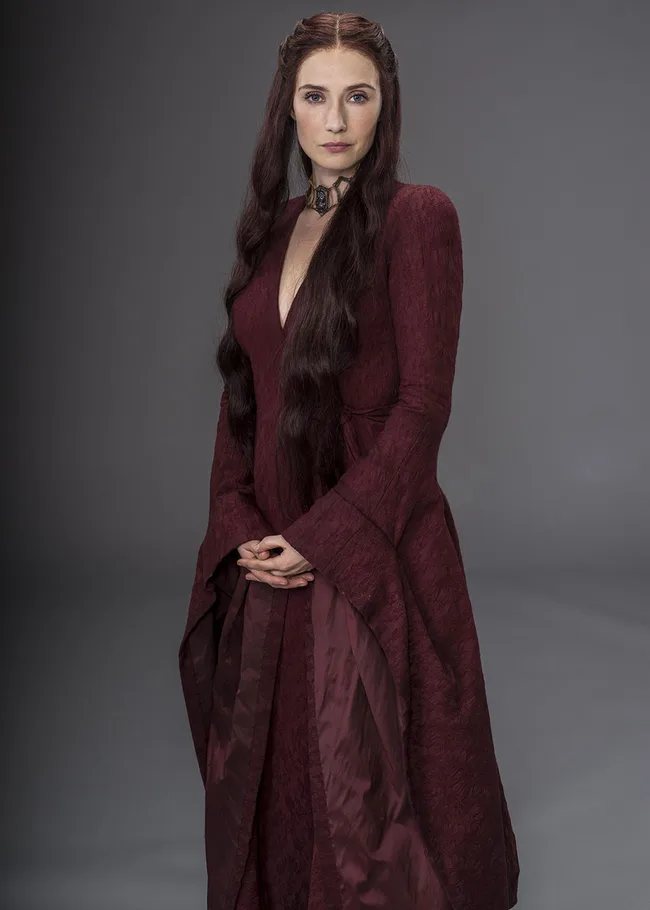 A woman in a long red gown stands with hands clasped, wearing a choker necklace, against a gray background.