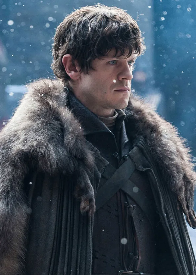 A person in a fur cloak stands in falling snow, looking serious.