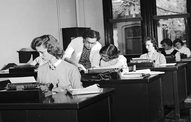 Classroom with women typing on typewriters, instructor assisting a student.