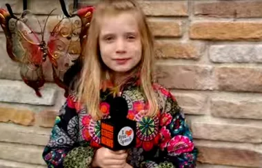 Young girl in a colorful coat holds a microphone, standing by a brick wall with butterfly decor.