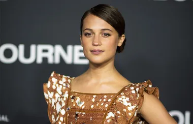 A person in a brown patterned dress poses against a dark background with text partially visible.