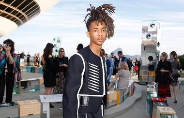 Young person with dreadlocks wearing a stylish black outfit at an outdoor event with people and modern decor in the background.