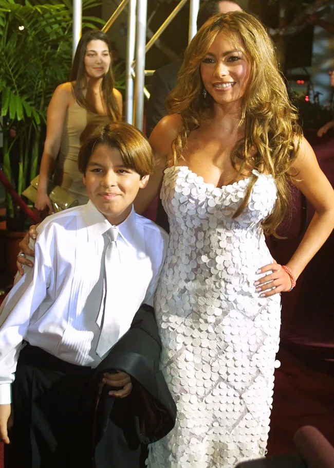 Woman in a sequined white dress poses with a young boy in a white shirt and tie at an event.