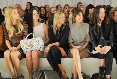 Front row at fashion show with five women seated, surrounded by audience members.