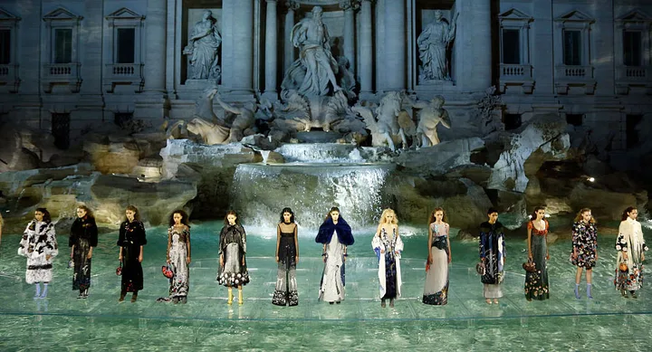 Fashion models in elegant dresses stand before Trevi Fountain, Rome, during a night runway show.